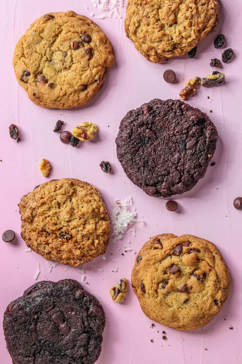 Assorted gourmet cookies including chocolate chip, oatmeal raisin, and dark chocolate varieties styled on a pink background with scattered nuts and chocolate pieces, perfect for a bakery dessert spread.