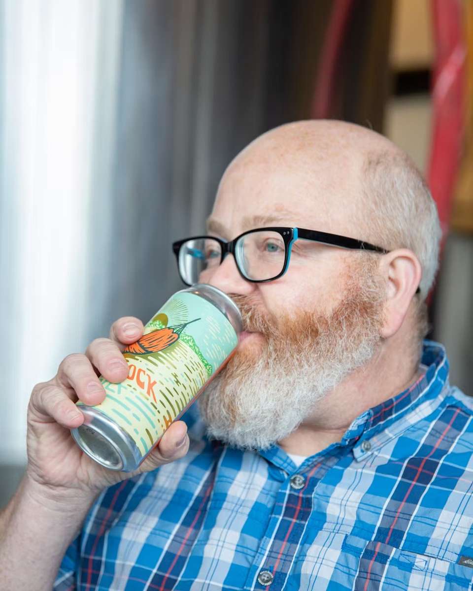 Man in glasses and plaid shirt drinks from a colorful can labeled "Hammock" with a stylized peach illustration.