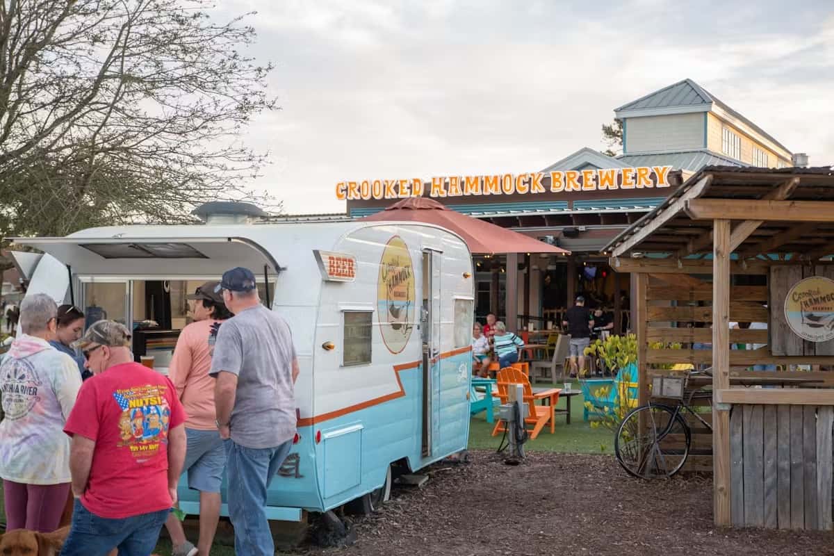 People line up at a retro camper trailer serving drinks outside Crooked Hammock Brewery with colorful seating and signage.