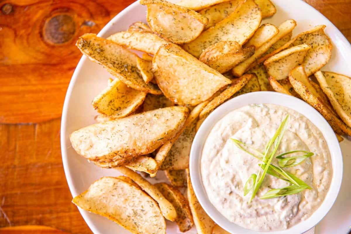 Crispy herb-seasoned potato wedges served with creamy onion dip topped with scallions on a white plate.