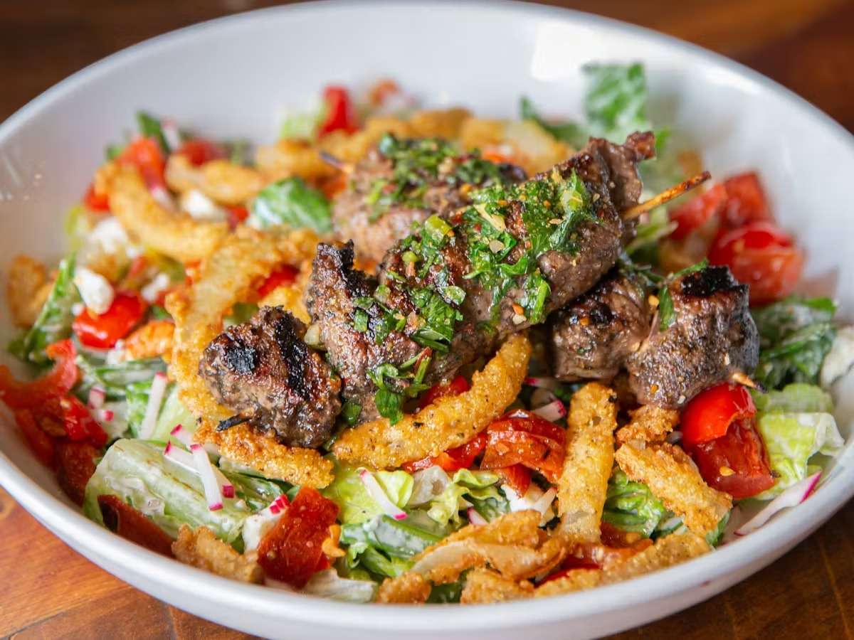 Grilled steak kabobs served over a fresh salad with crispy onions, tomatoes, lettuce, and herbs in a white bowl.
