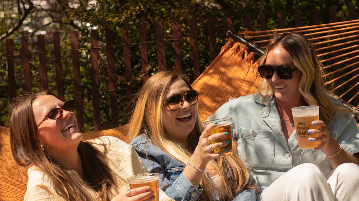 Three women in sunglasses laugh and toast beers while lounging together on an orange hammock outdoors.
