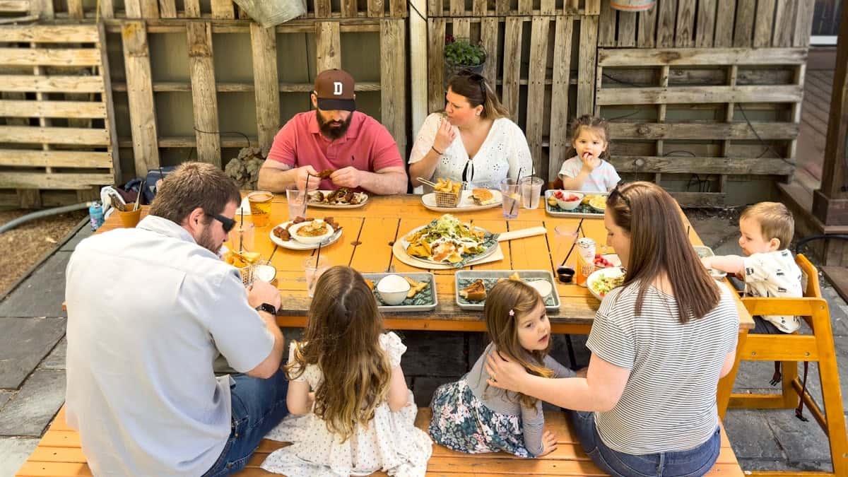 Large family enjoying an outdoor meal together around a wooden picnic table with various plates of food and drinks.