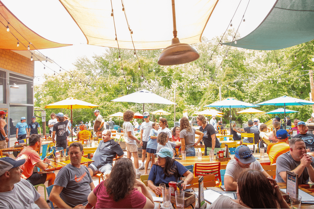 Outdoor dining area with colorful umbrellas, crowded tables, and people socializing under string lights on a sunny day.