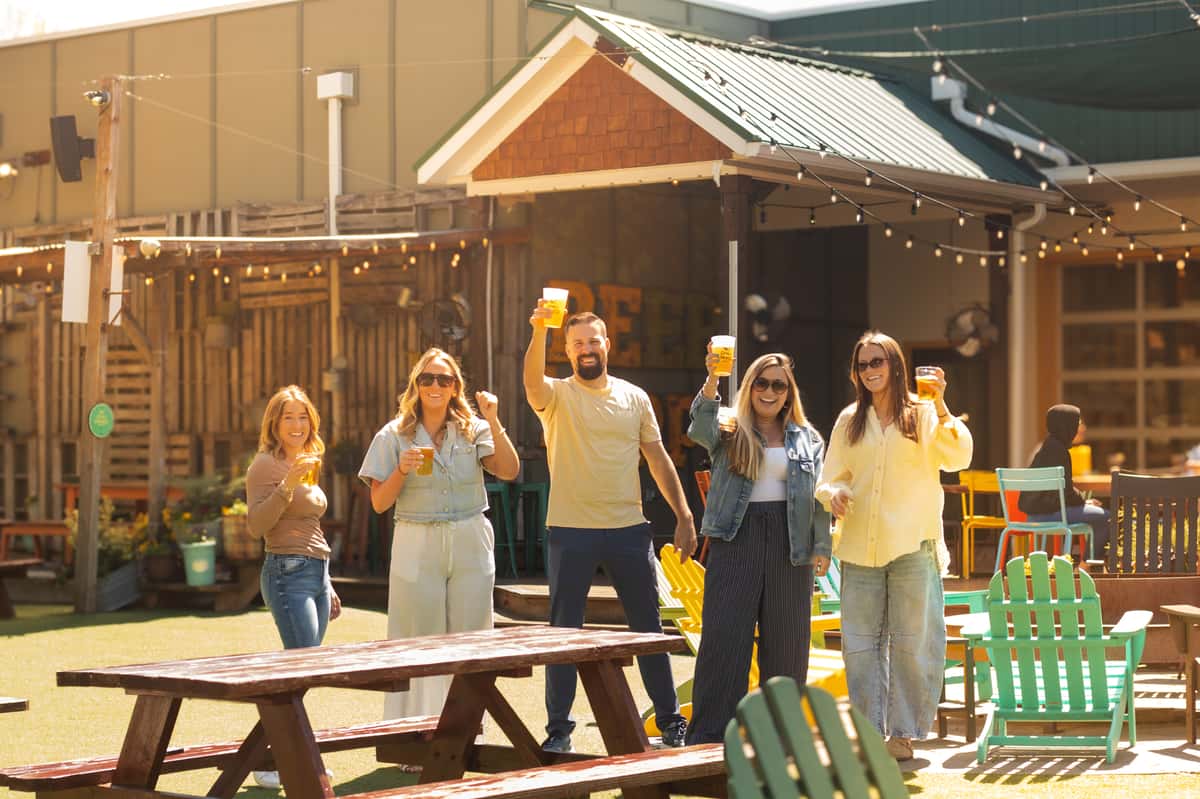 People raising glasses of beer while smiling in a sunny outdoor patio with picnic tables and colorful chairs.