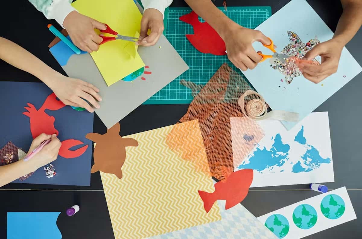 Children's hands crafting sea animals with colorful paper, scissors, and glitter on a table covered in creative materials.
