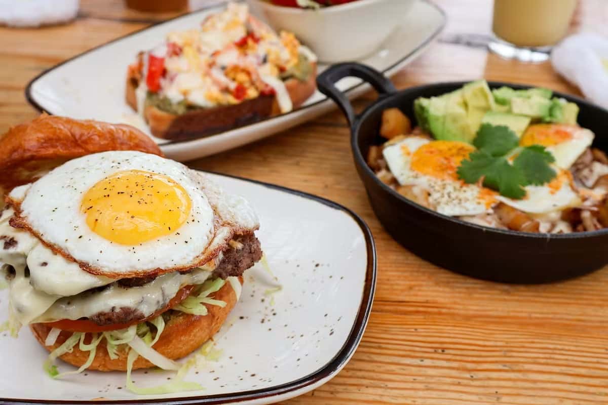 Burger with fried egg, cheesy skillet hash with avocado, and loaded toast on a wooden table.
