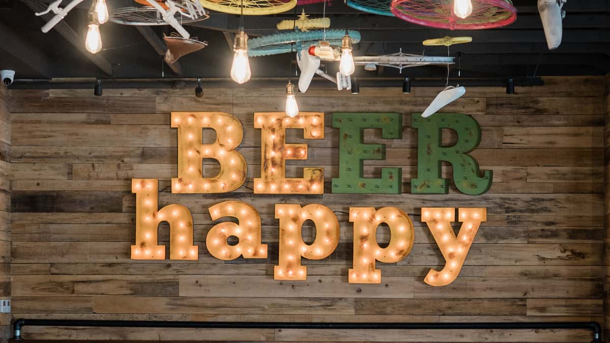 Rustic wooden wall with illuminated marquee letters spelling "BEER happy" under exposed light bulbs and hanging decorative planes.