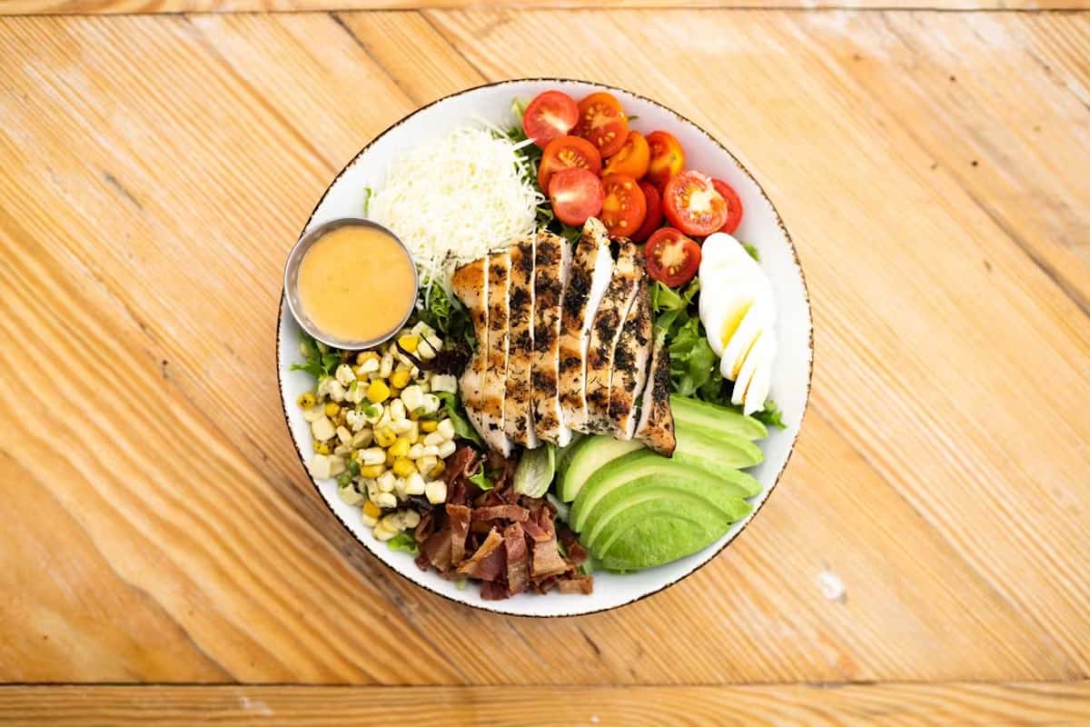 Grilled chicken Cobb salad with cherry tomatoes, avocado, egg, bacon, corn, shredded cheese, and dressing on a wooden table.