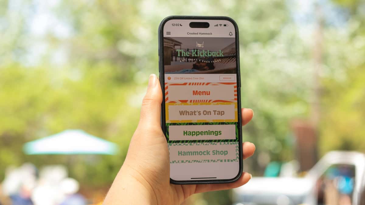 Person holding a smartphone displaying The Kickback menu from Crooked Hammock, with outdoor seating blurred in the background.