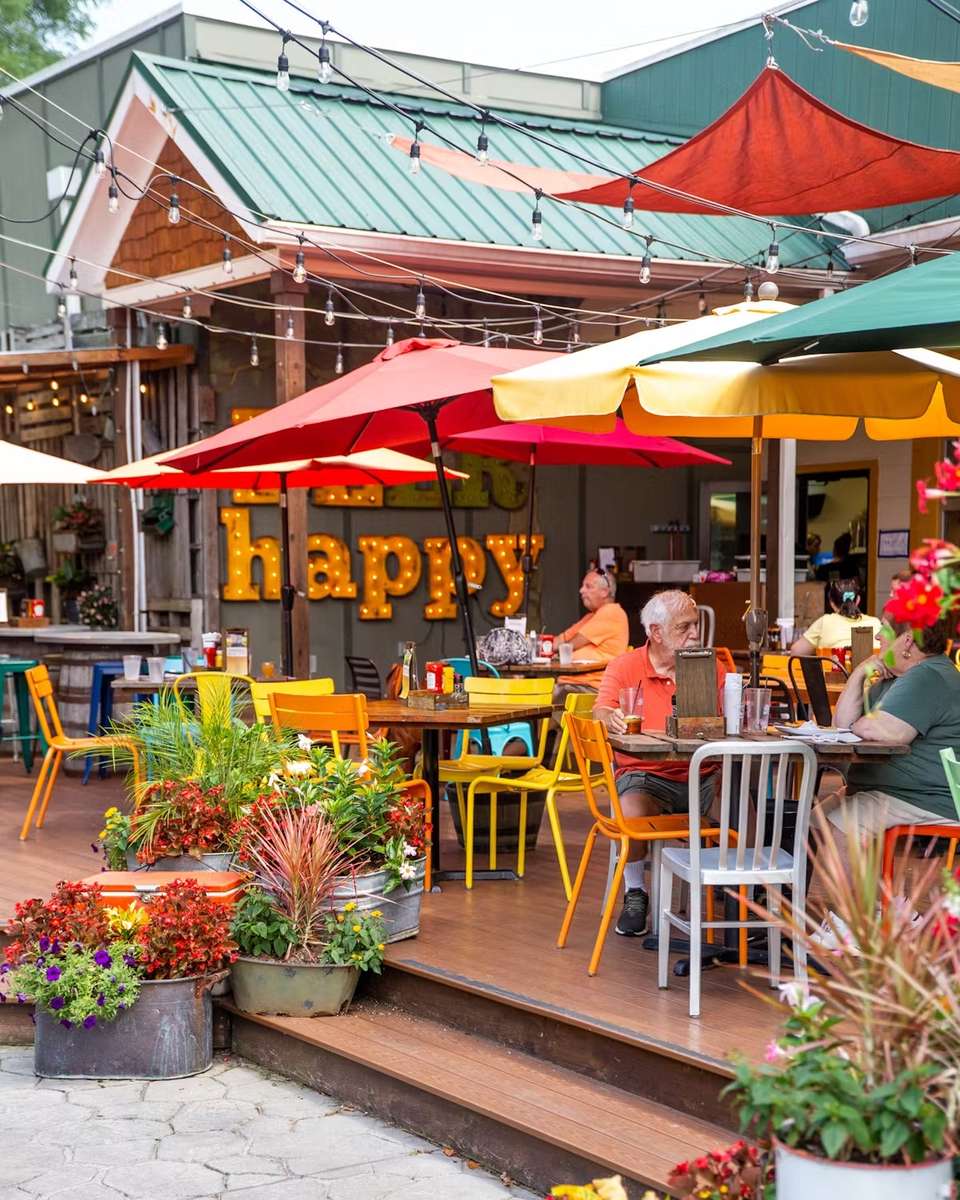 Colorful outdoor patio at Crooked Hammock Brewery with guests dining under umbrellas and a large lit "happy" sign in the background.
