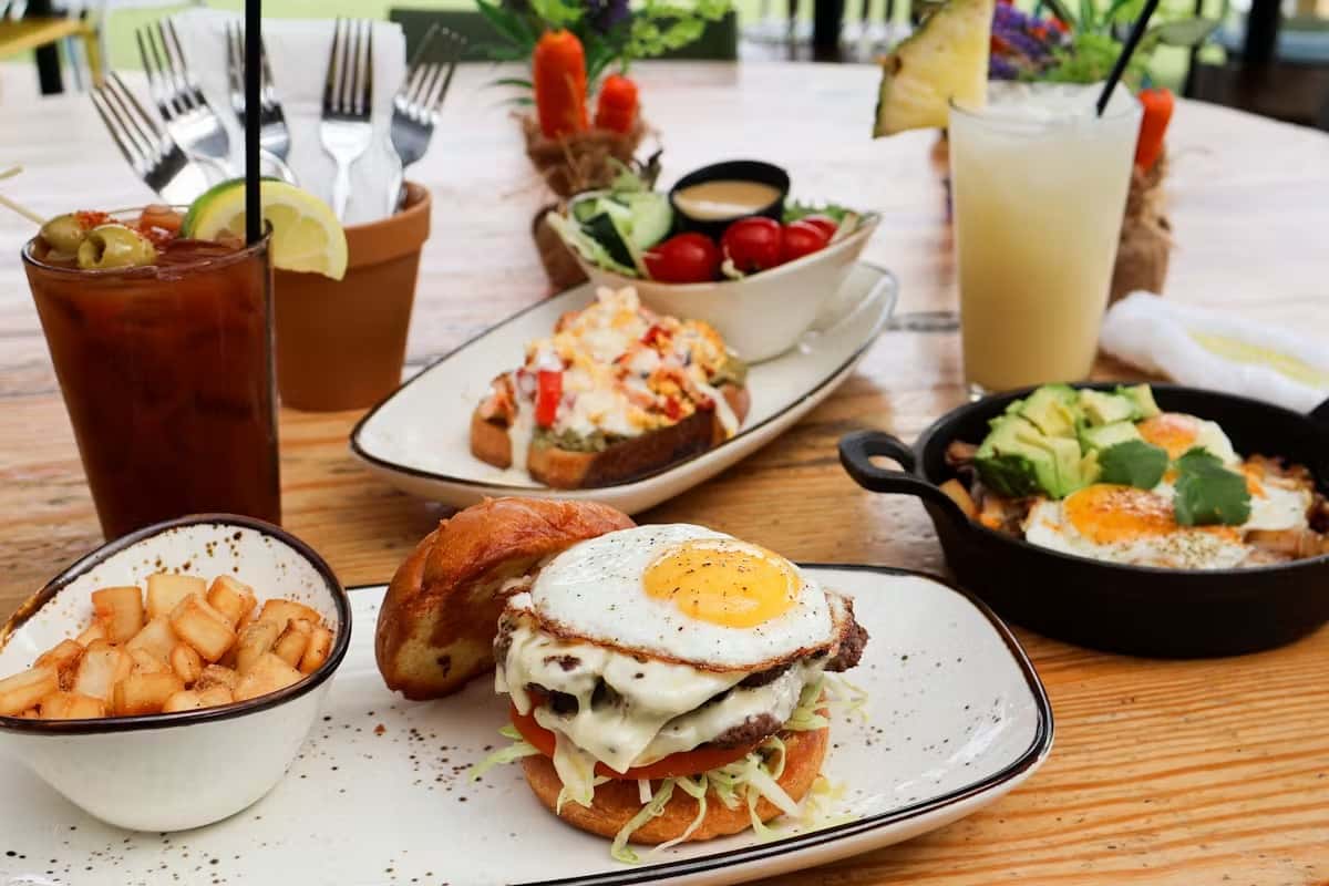 A breakfast burger with a fried egg is served alongside seasoned potatoes, with cocktails and other brunch dishes on a wood table.