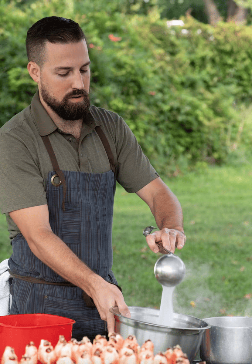 Man in an apron ladles steaming liquid into a bowl at an outdoor food setup with shrimp neatly arranged in front.