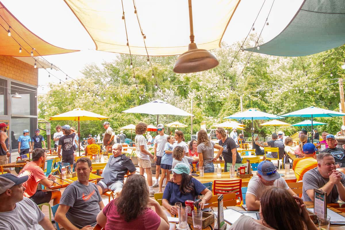 Crowd enjoys food and drinks on a colorful patio under umbrellas and shade sails at Crooked Hammock Brewery.
