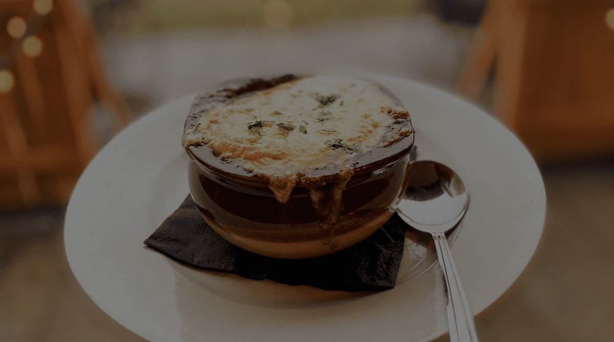 French onion soup in a brown bowl with melted cheese and parsley, on a white plate with a spoon.