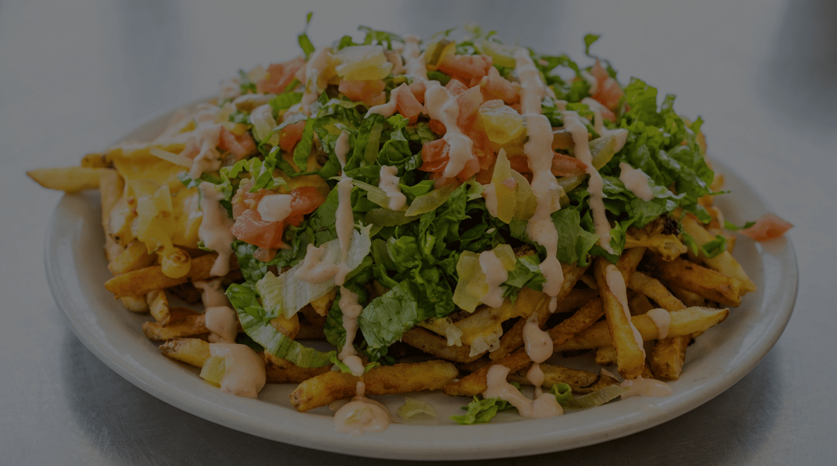 Loaded fries with melted cheese, chopped lettuce, diced tomatoes, pickles, and a drizzle of pink sauce on a white plate.