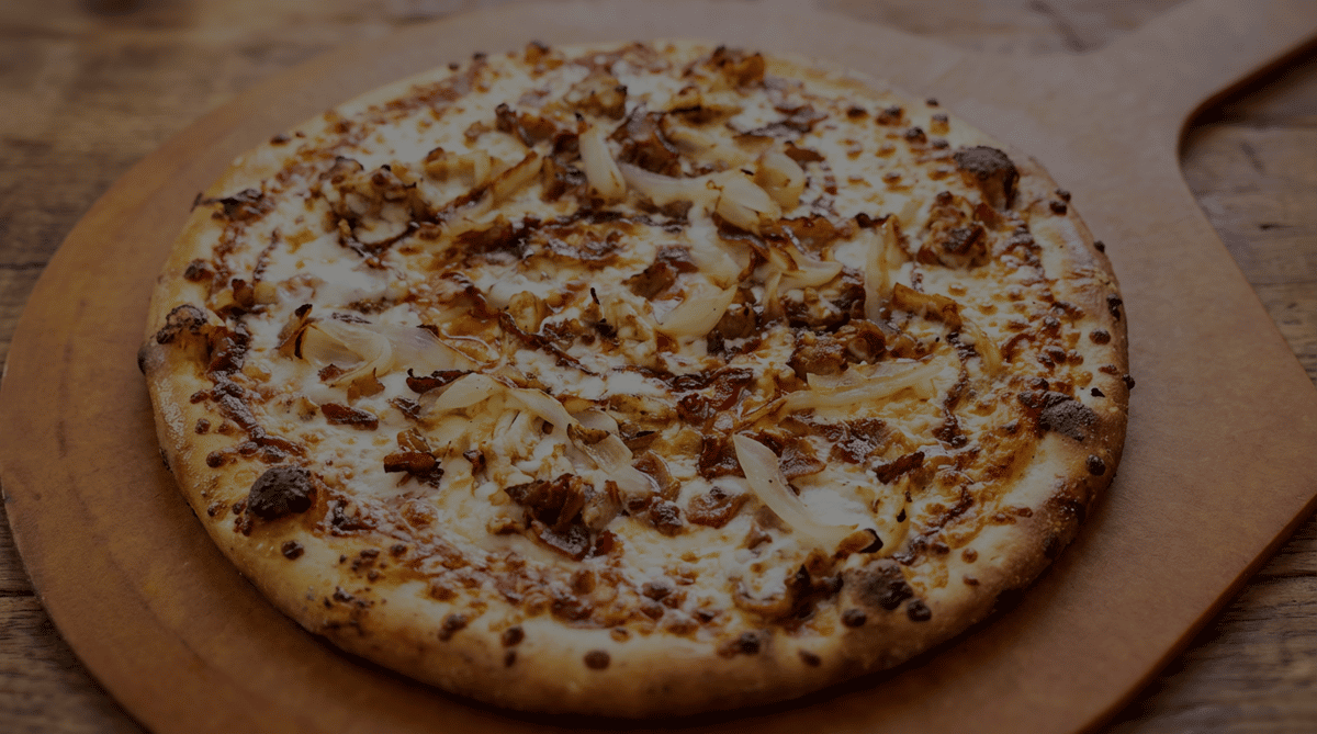 Round pizza with melted cheese, caramelized onions, and chopped meat on a wooden peel.