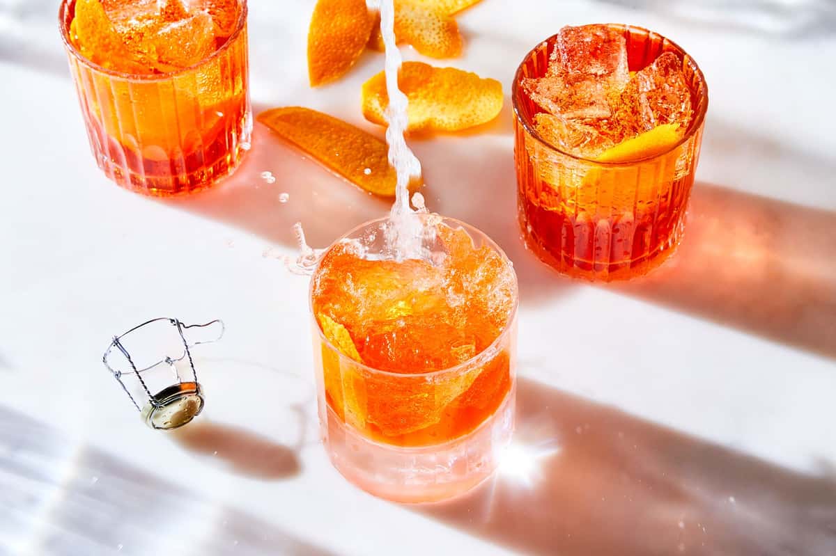 Bright orange spritz cocktails with ice and citrus peel being poured on a sunlit table