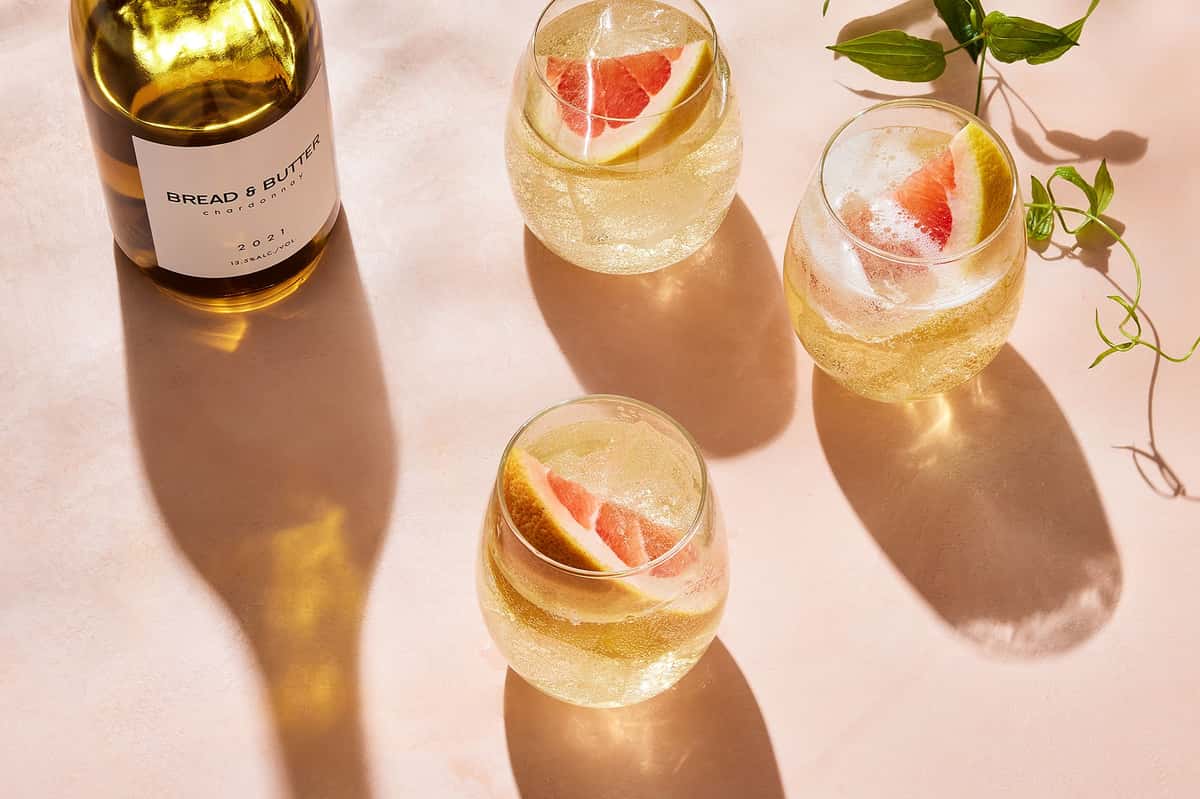 Chilled white wine spritzers with grapefruit slices in stemless glasses, served beside a bottle of chardonnay on a sunlit table.