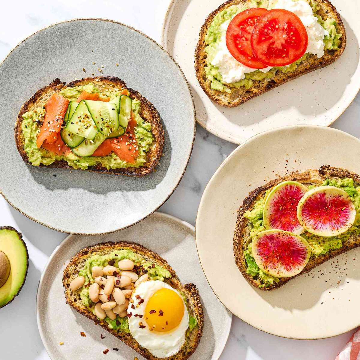 Assorted avocado toast variations on rustic bread, topped with smoked salmon and cucumber, tomato and ricotta, watermelon radish slices, and a fried egg with white beans, served on ceramic plates.