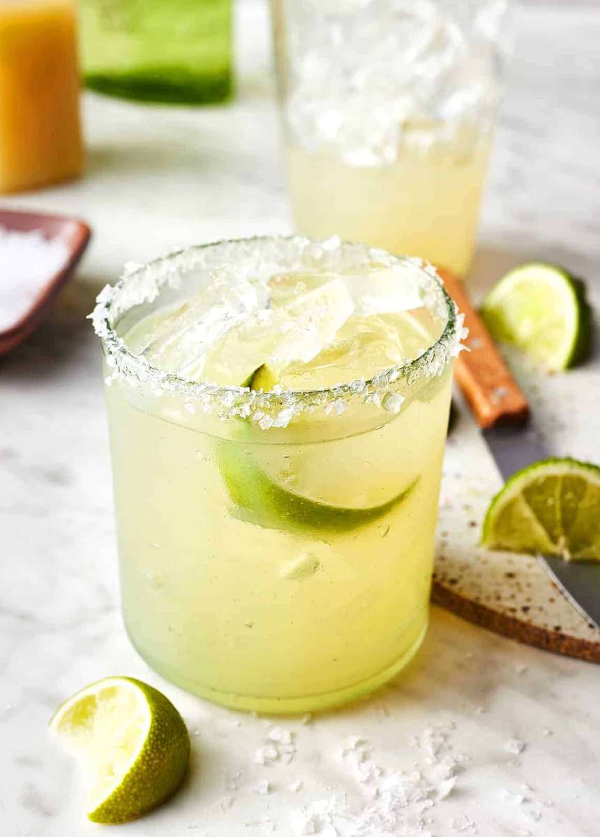 Fresh lime margarita with salted rim, served over ice with lime wedges on a light stone surface.
