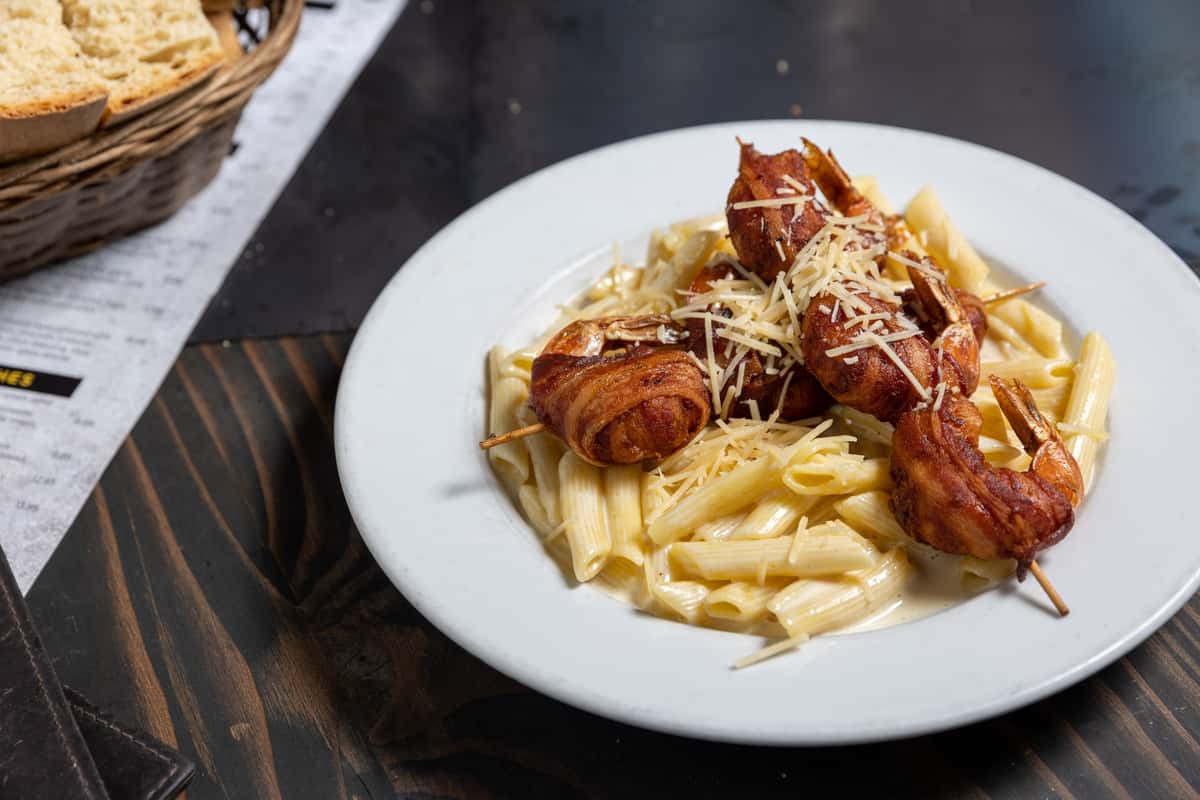 Creamy penne pasta topped with skewered bacon-wrapped shrimp and shredded Parmesan cheese on a white plate.