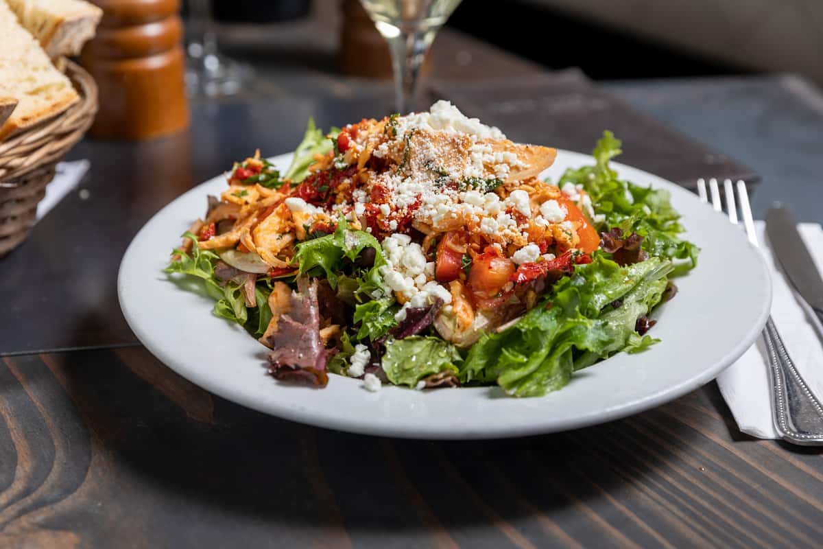 Fresh mixed greens salad topped with grilled chicken, diced tomatoes, crumbled feta cheese, and shredded carrots on a white plate.