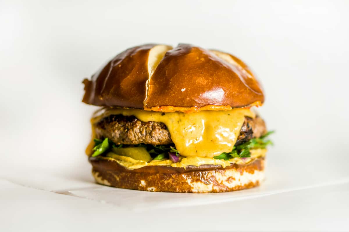 Juicy cheeseburger on a soft pretzel bun with melted cheddar, pickles, and house sauce on a clean white background.