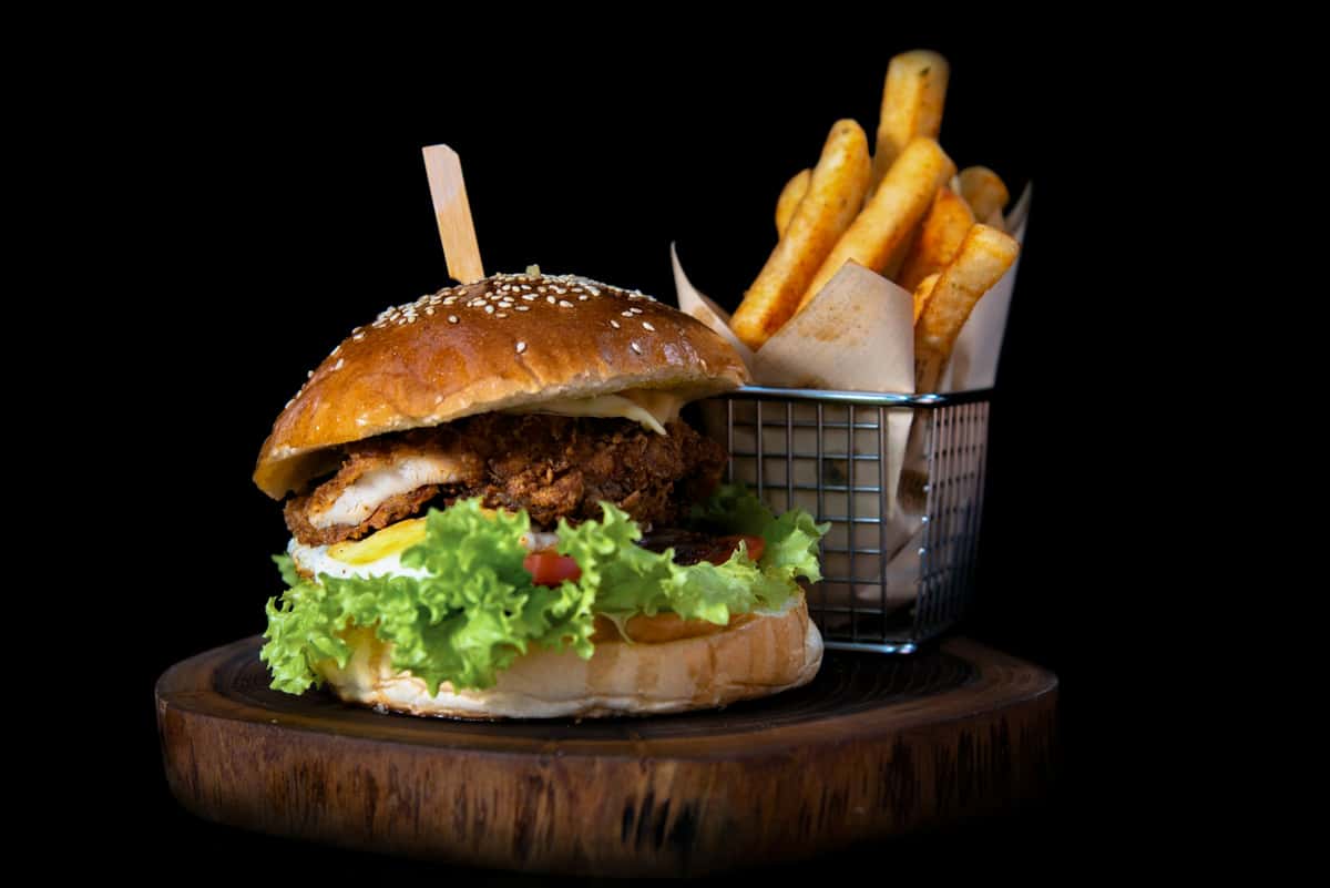 **Alt text:** Crispy fried chicken burger with fresh lettuce and sauce on a sesame brioche bun, served with thick-cut golden fries in a metal basket on a wooden board against a dark background.