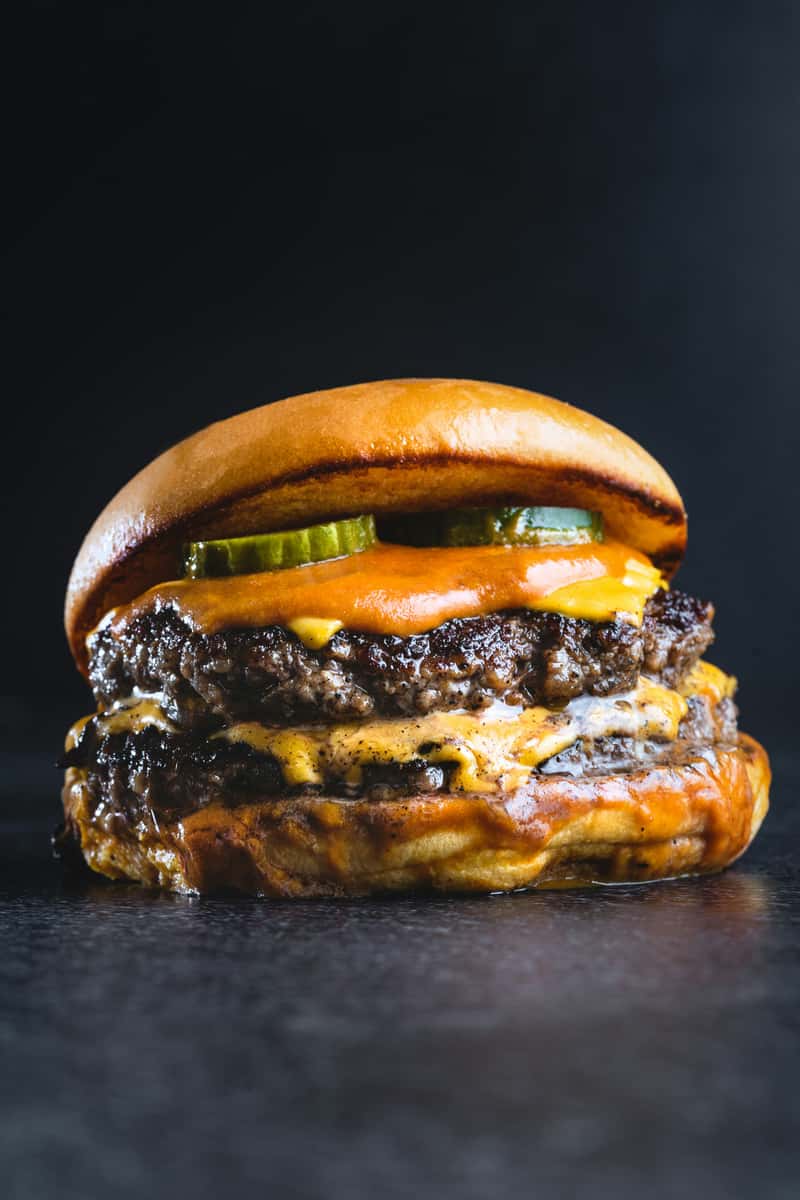 Double smash cheeseburger with melted cheese, pickles, and tangy burger sauce on a toasted brioche bun against a dark background.