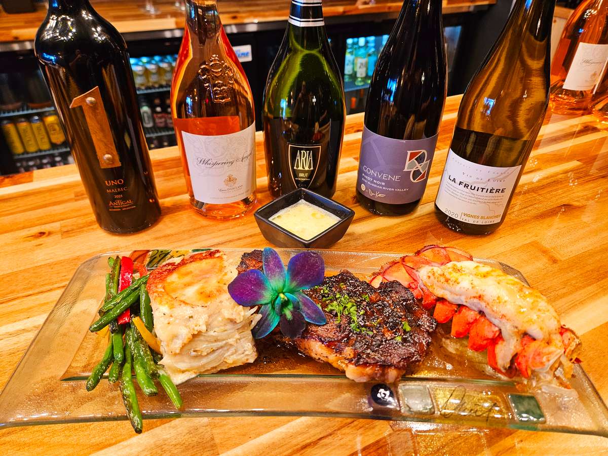 Surf and turf entrée featuring grilled steak, lobster tail, scalloped potatoes, and sautéed vegetables, elegantly plated with a purple edible flower garnish, served alongside a flight of fine wines on a wooden bar counter.