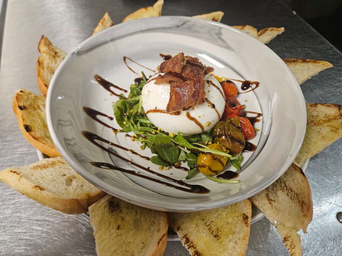 Fresh burrata cheese served over arugula, roasted cherry tomatoes, and pesto, topped with crispy bacon and drizzled with balsamic glaze, surrounded by grilled crostini on a white plate.