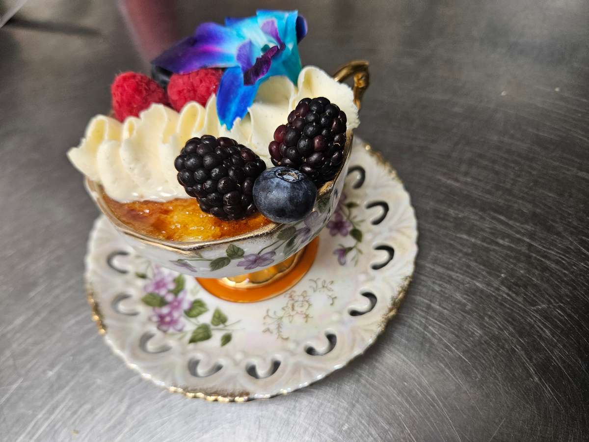 Crème brûlée served in an ornate floral teacup, topped with whipped cream, fresh blackberries, raspberries, a blueberry, and a vibrant blue orchid, placed on a matching vintage saucer.
