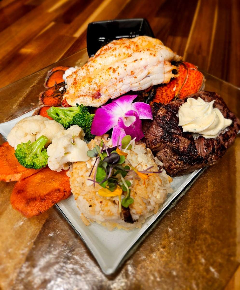 Gourmet surf and turf plate featuring a broiled lobster tail, grilled filet mignon topped with herbed butter, vegetable medley, seasoned rice, and sweet potato slices, garnished with an orchid flower and microgreens, served on a glass plate over a wooden table.