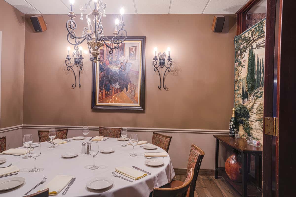 Private dining room with round table set in white linens, wall sconces, framed art, and chandelier lighting