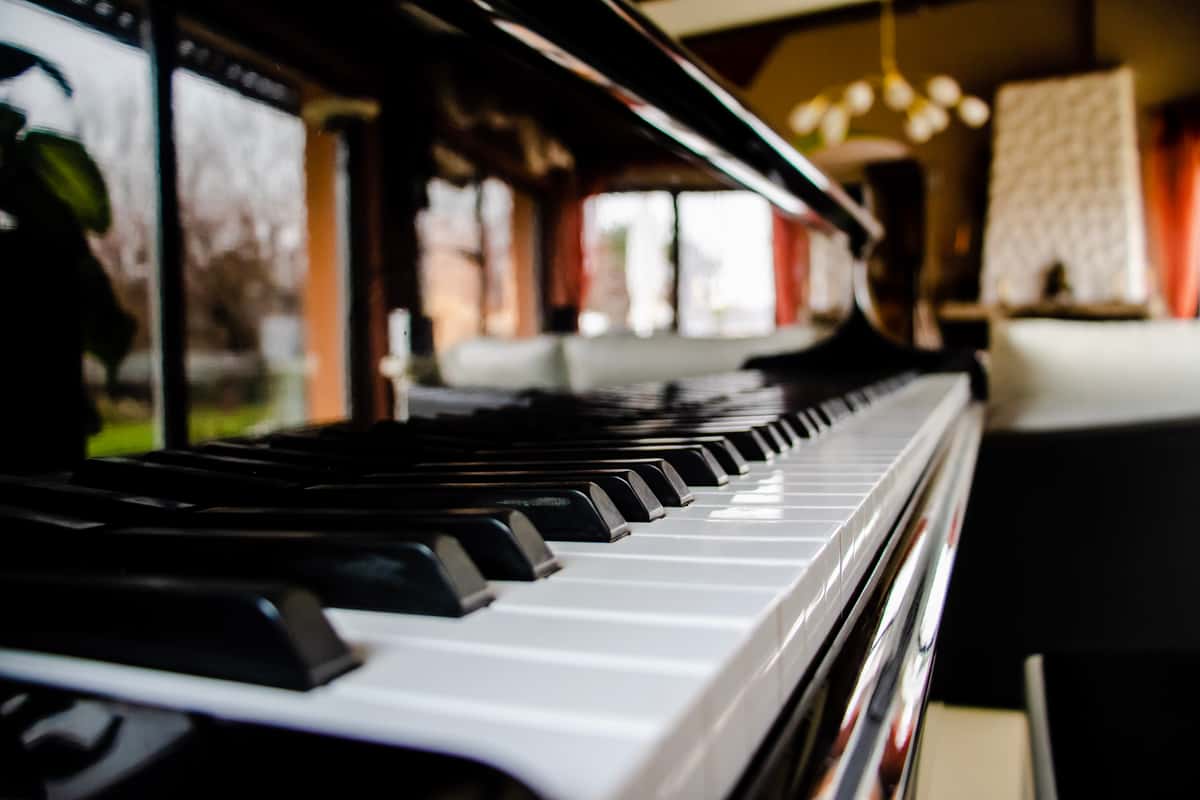 A low-angle, close-up view of the keys of a black grand piano, with a blurred dining room visible in the background.