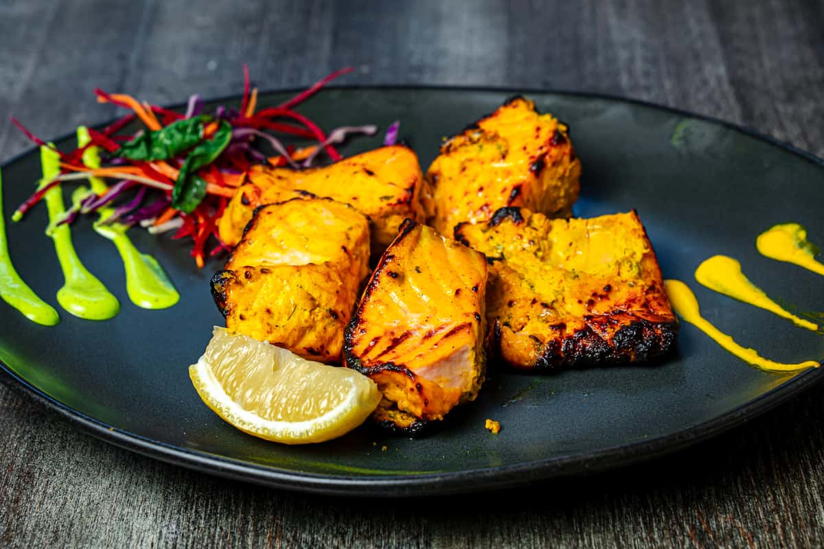 Grilled chicken tikka cubes marinated in spices, served with lemon wedge, salad garnish, and sauces on a black plate.