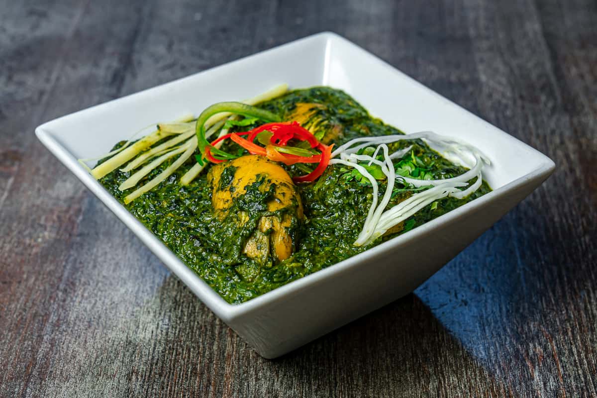 Palak chicken curry with tender chicken pieces in a rich spinach-based Indian gravy, garnished with julienned ginger, fresh herbs, and sliced chilies in a square white bowl.