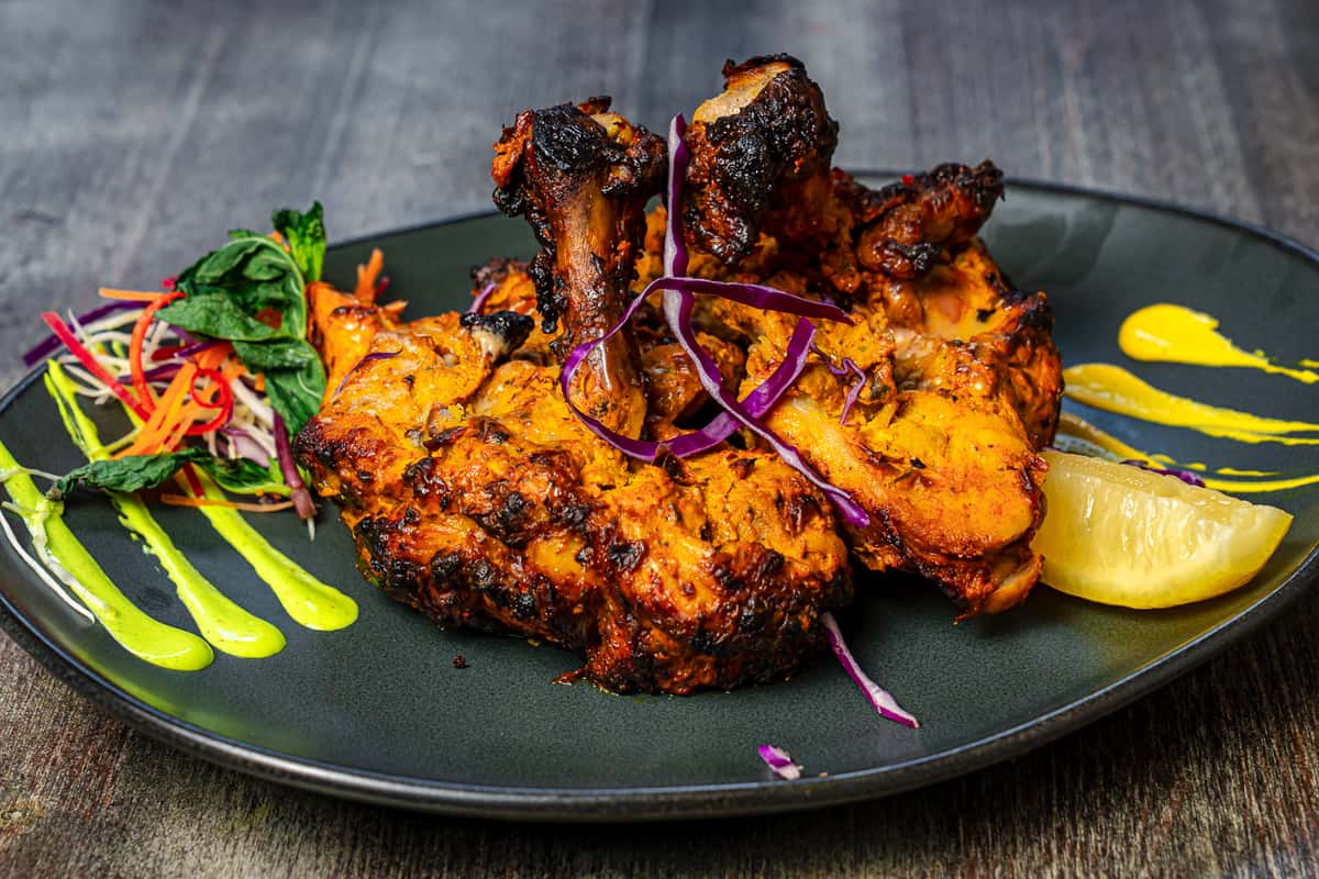 Tandoori grilled chicken drumsticks with charred edges, served with lemon wedge, fresh salad garnish, and vibrant sauce on a black plate.