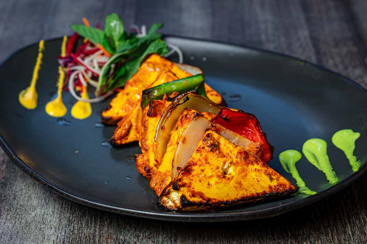 Grilled tandoori-style paneer with roasted bell peppers and onions, served on a matte black plate with colorful chutney sauces and a fresh vegetable salad garnish—vibrant Indian restaurant appetizer presentation.