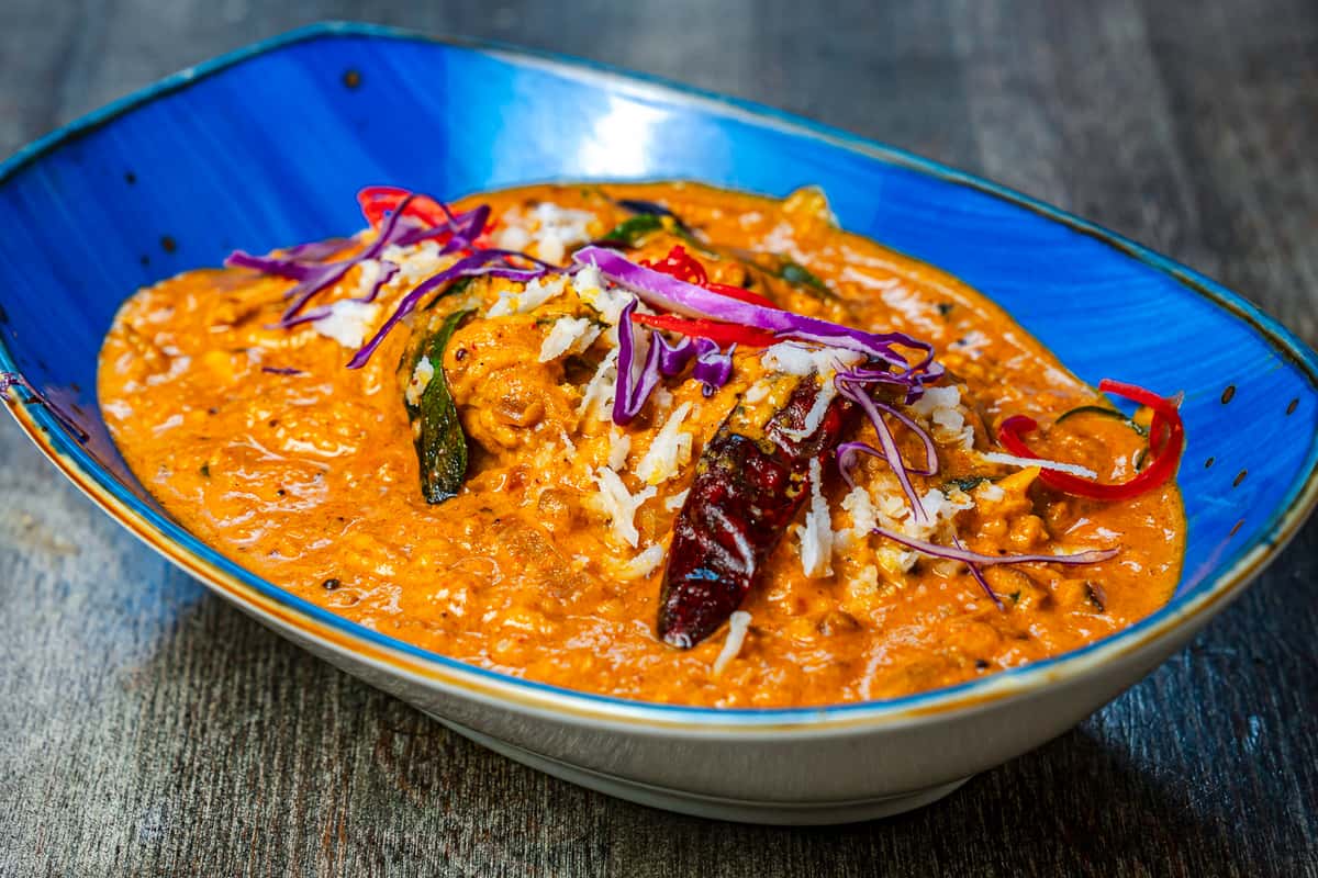 Creamy South Indian–style curry with tender protein in a rich coconut-based sauce, garnished with dried red chilies, curry leaves, shredded coconut, and fresh cabbage in a vibrant blue restaurant bowl.