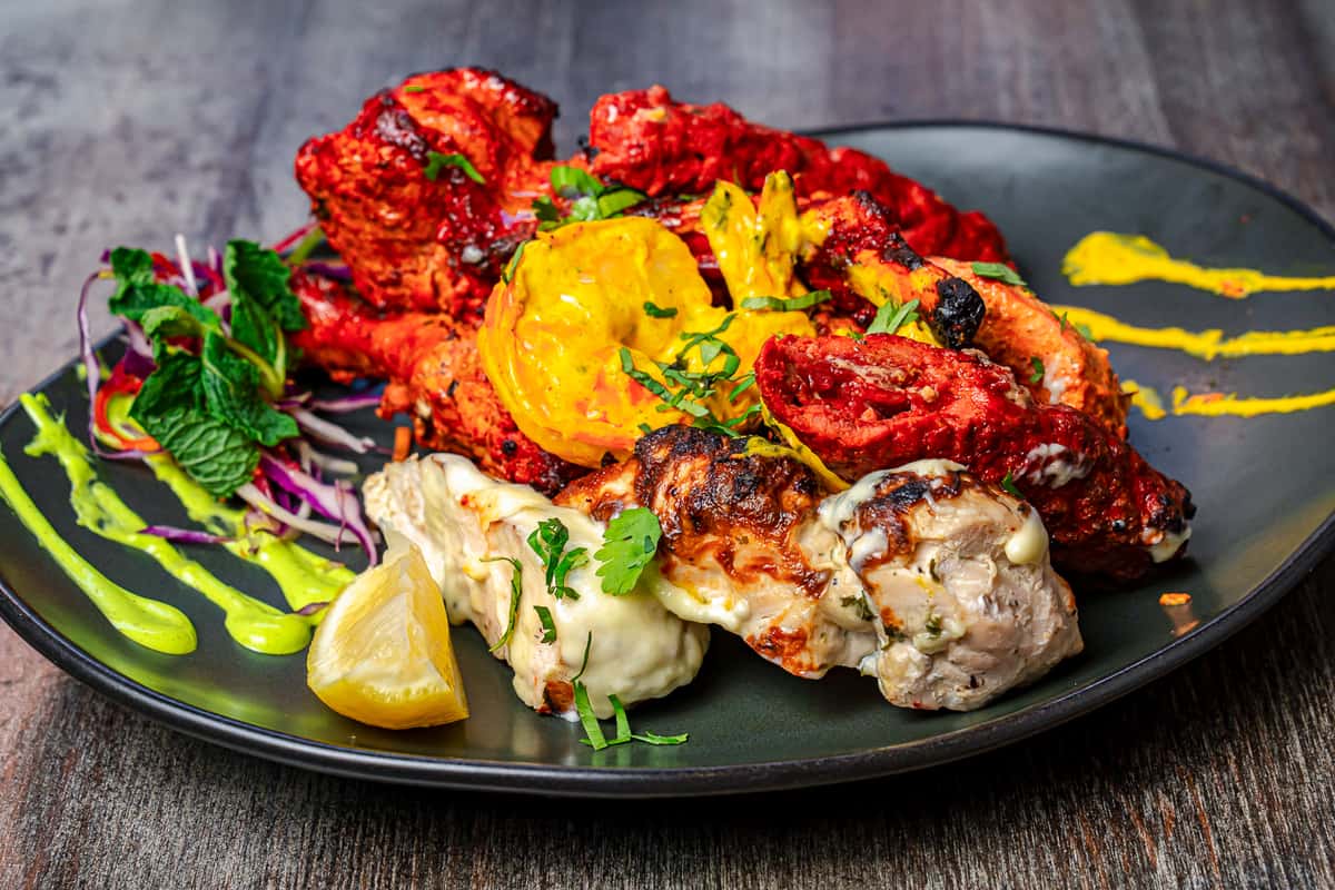 Indian mixed tandoori grill platter with chicken tikka, malai chicken, and seekh kebab, served with roasted vegetables, lemon wedge, fresh herbs, and vibrant mint and turmeric chutney on a modern black restaurant plate.