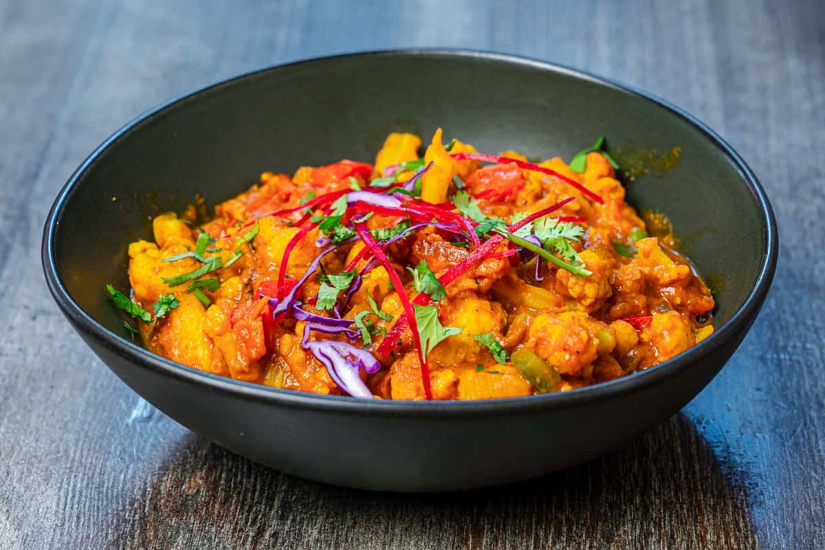 Indian vegetable curry with mixed vegetables simmered in a rich spiced tomato gravy, garnished with fresh cilantro, shredded cabbage, and herbs in a modern black bowl.
