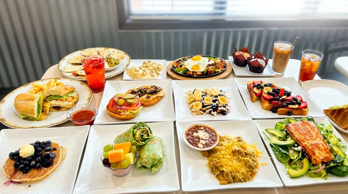 A child poses enthusiastically at a restaurant table laden with a variety of breakfast and lunch dishes.