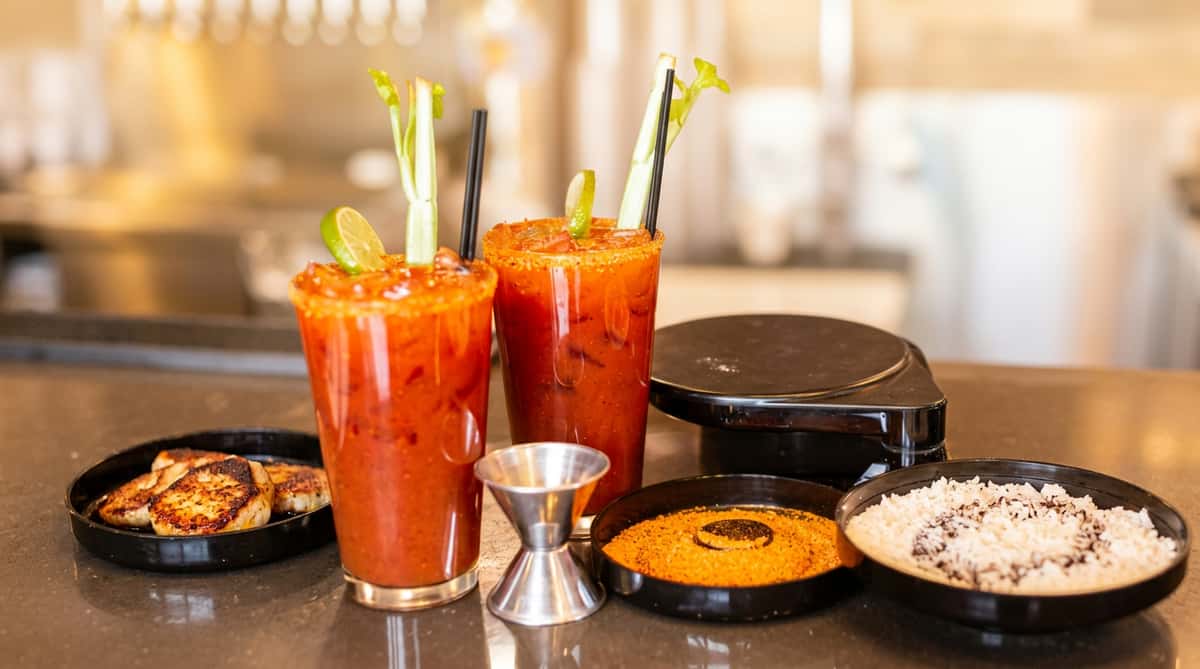 Two Bloody Mary cocktails with lime wedges, celery stalks, and spiced rims, alongside ingredients.