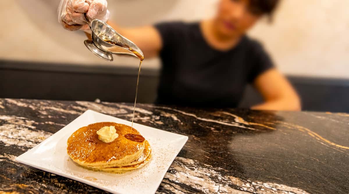 Pancakes with butter and powdered sugar on a white plate, with syrup being poured from a gravy boat.