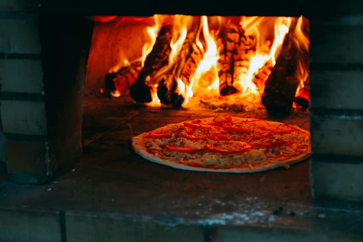 Authentic wood-fired pizza baking in a brick oven, with flames and burning logs creating a rustic, smoky cooking atmosphere.