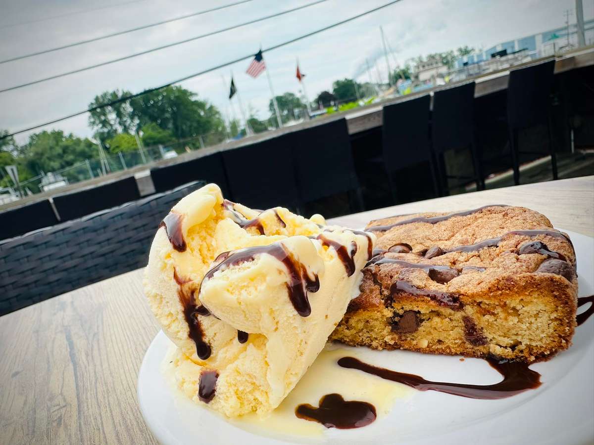 Vanilla ice cream topped with chocolate sauce, served beside a warm chocolate chip cookie bar on a white plate.