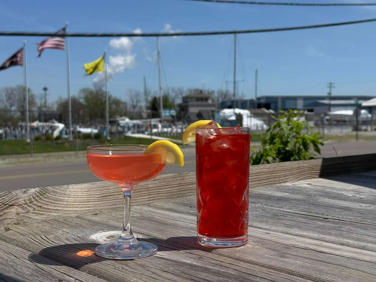 Two red cocktails with lemon garnishes sit on a wooden patio table with a marina and flags in the background.