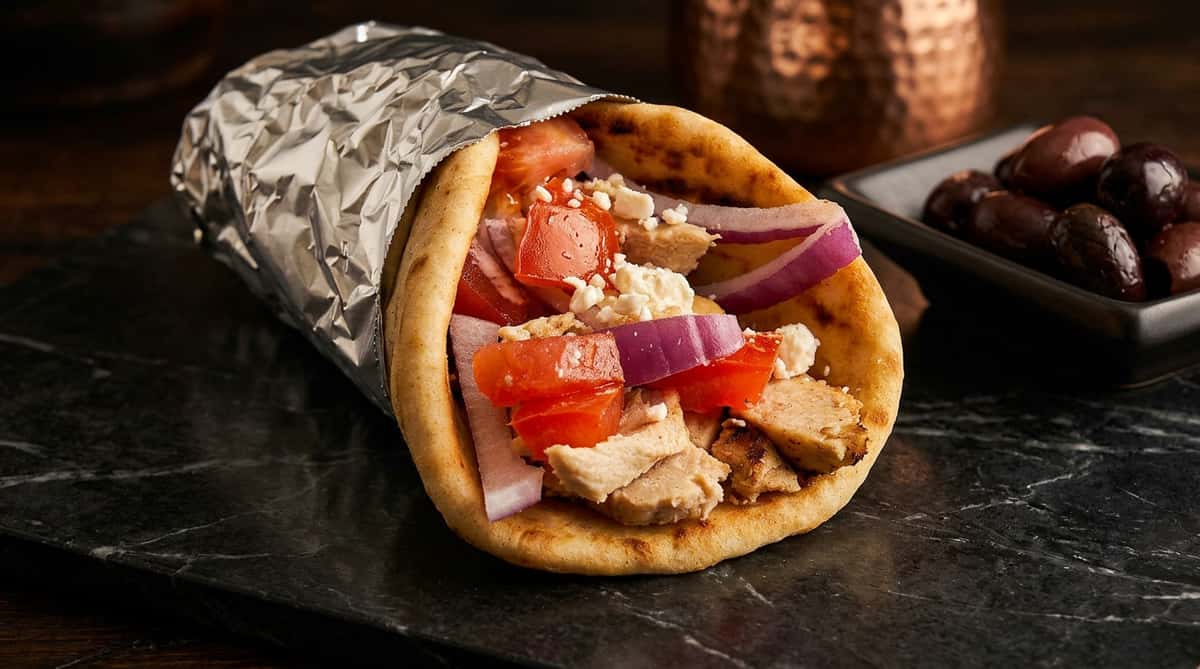 Chicken gyro in pita bread with red onions, tomatoes, feta cheese, and tzatziki sauce, partially wrapped in foil.